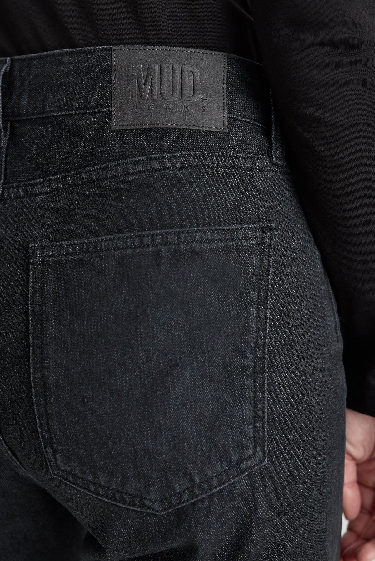 Close-up of MUD Jeans featuring sustainable dark gray denim, showcasing back pocket and leather label, worn with black top.