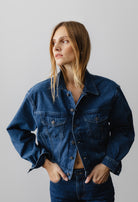 Casual denim jacket in deep indigo worn by a person with hands in pockets, featuring button details and a classic collar.