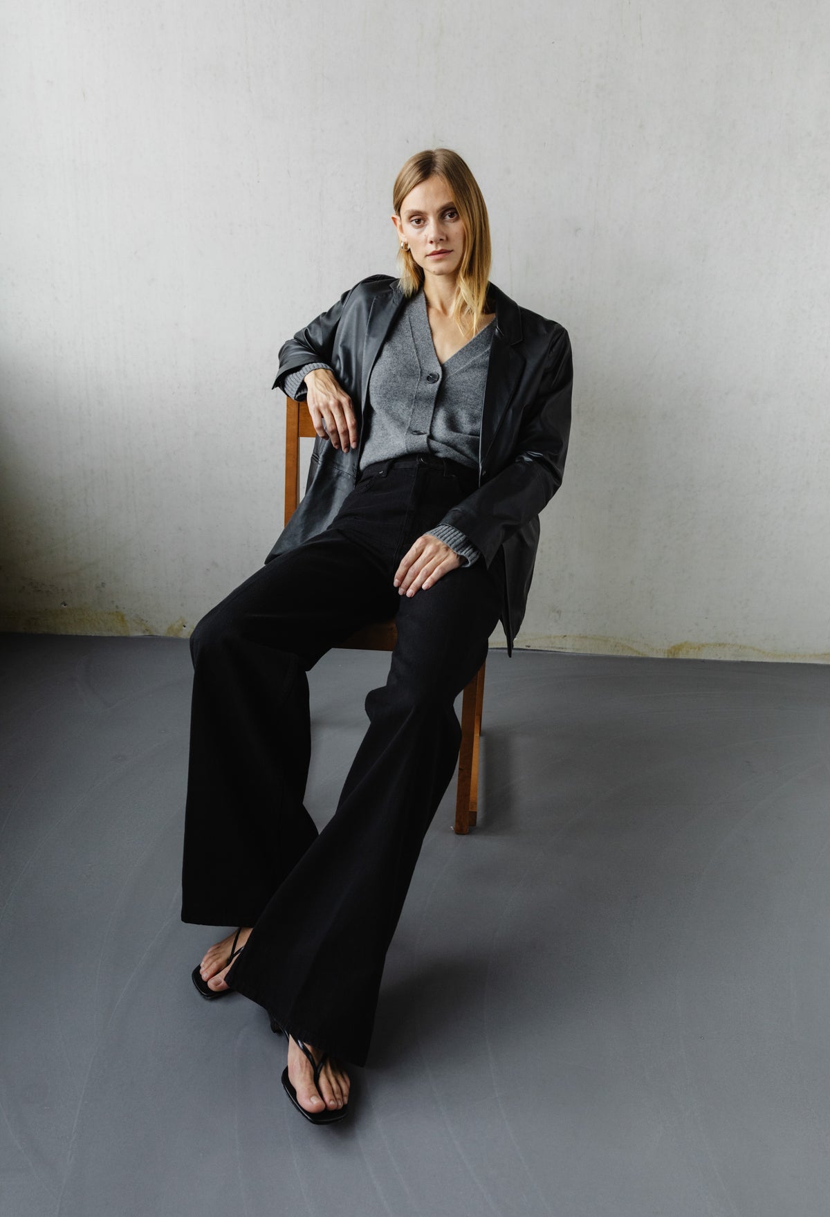 Stylish woman in a gray cardigan and black wide-leg pants, seated on a wooden chair against a minimalist concrete wall.