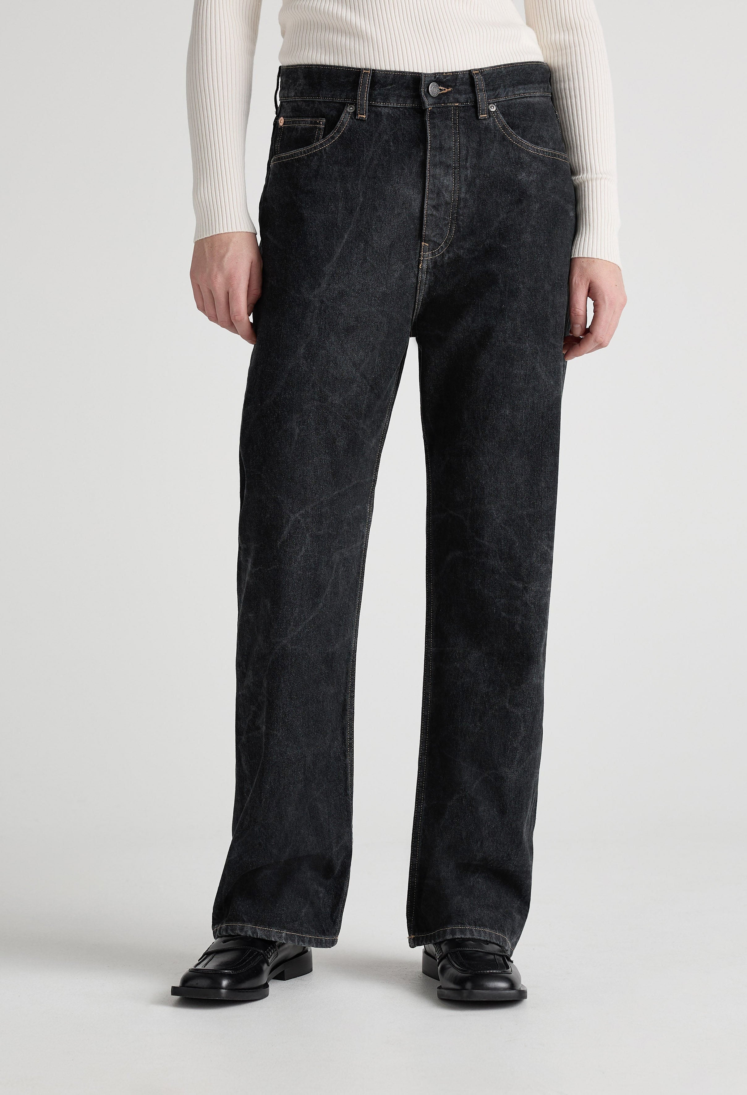 A model wearing loose-fitting black denim jeans with a ribbed, white long-sleeve top and black leather shoes.