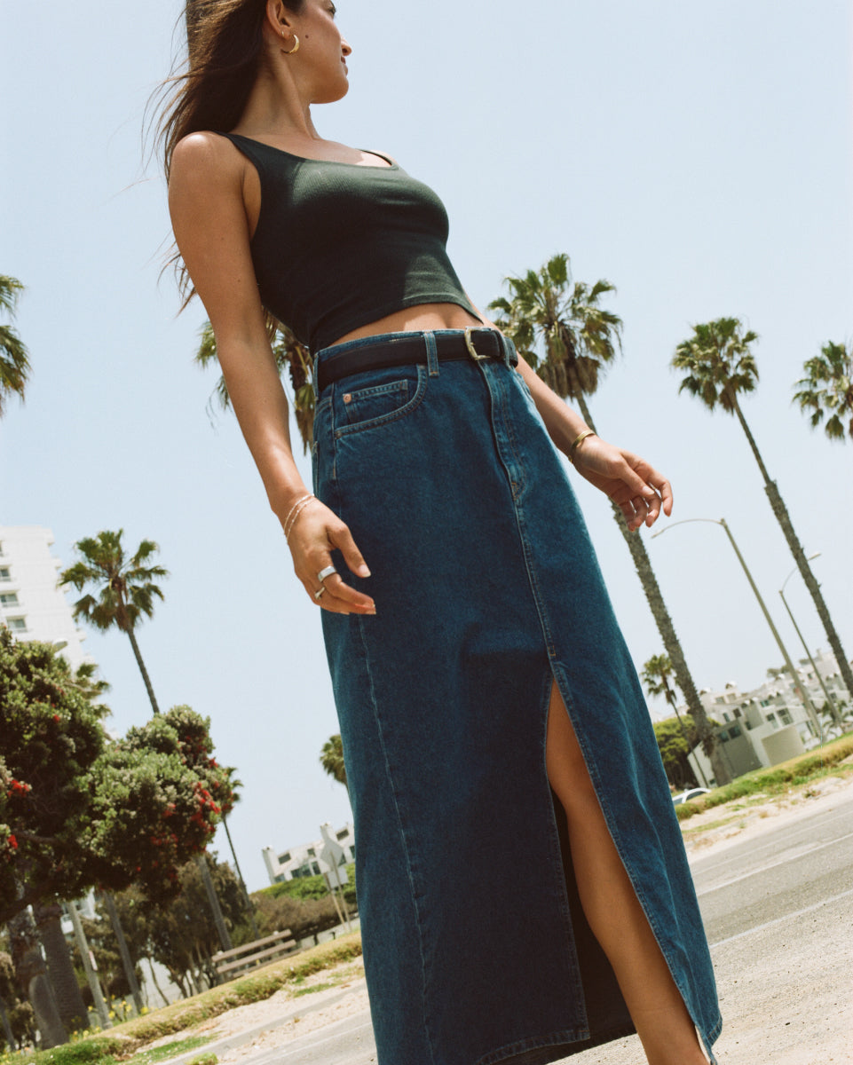 Woman in a sleeveless black top and long denim skirt with a side slit, standing outdoors under a clear sky.