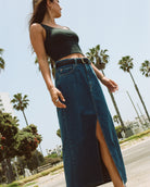 Woman in a sleeveless black top and long denim skirt with a side slit, standing outdoors under a clear sky.