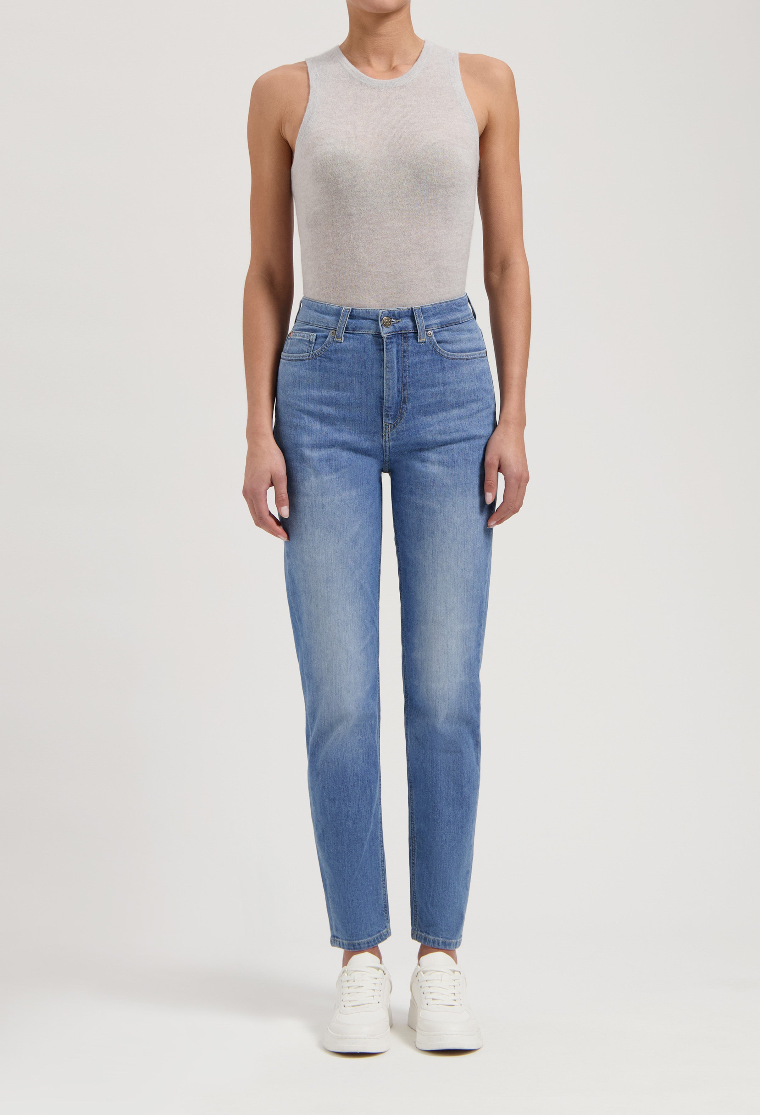 Woman wearing light gray sleeveless top and high-waisted blue jeans, paired with white sneakers, isolated on white background.