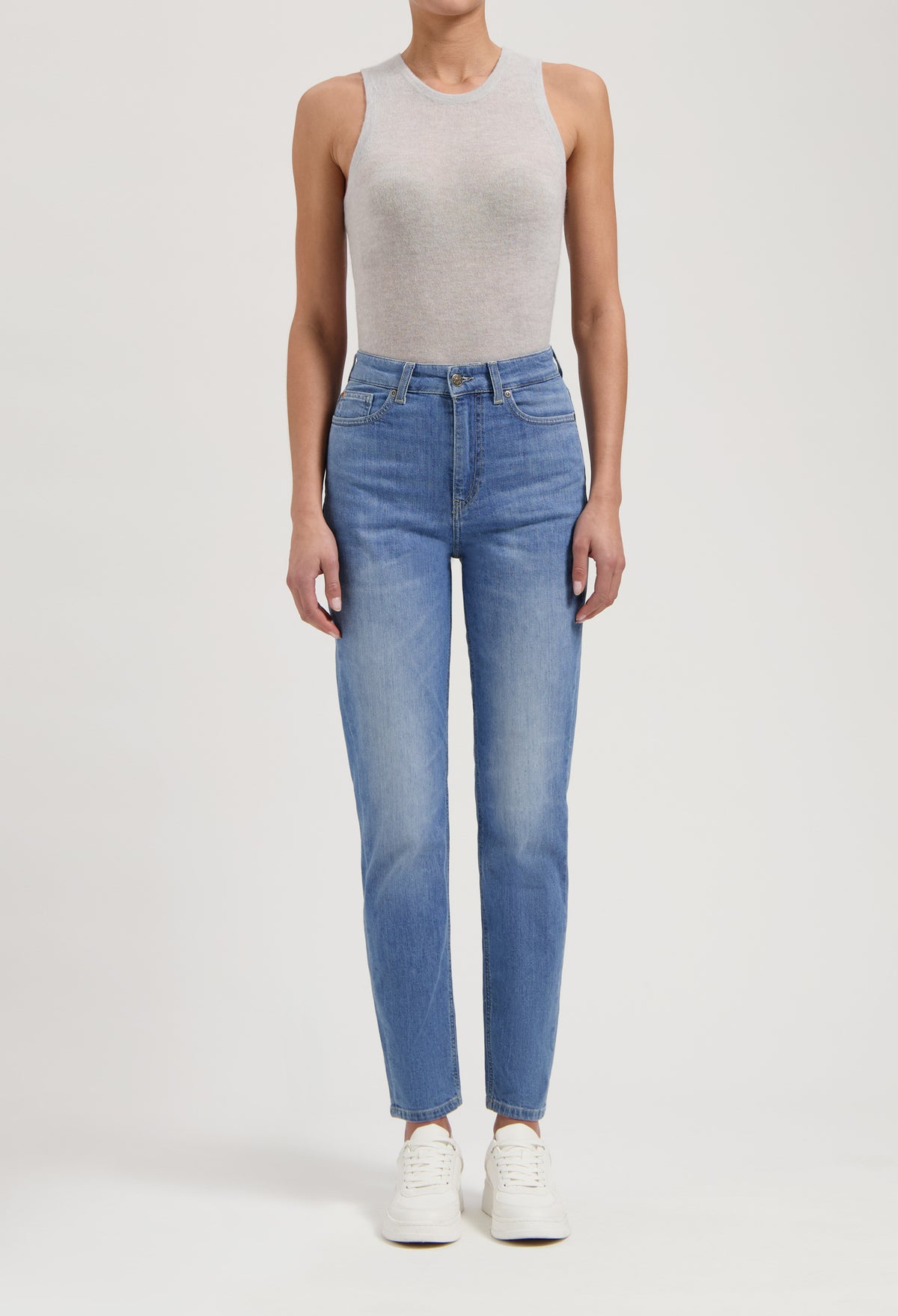 Woman wearing light gray sleeveless top and high-waisted blue jeans, paired with white sneakers, isolated on white background.