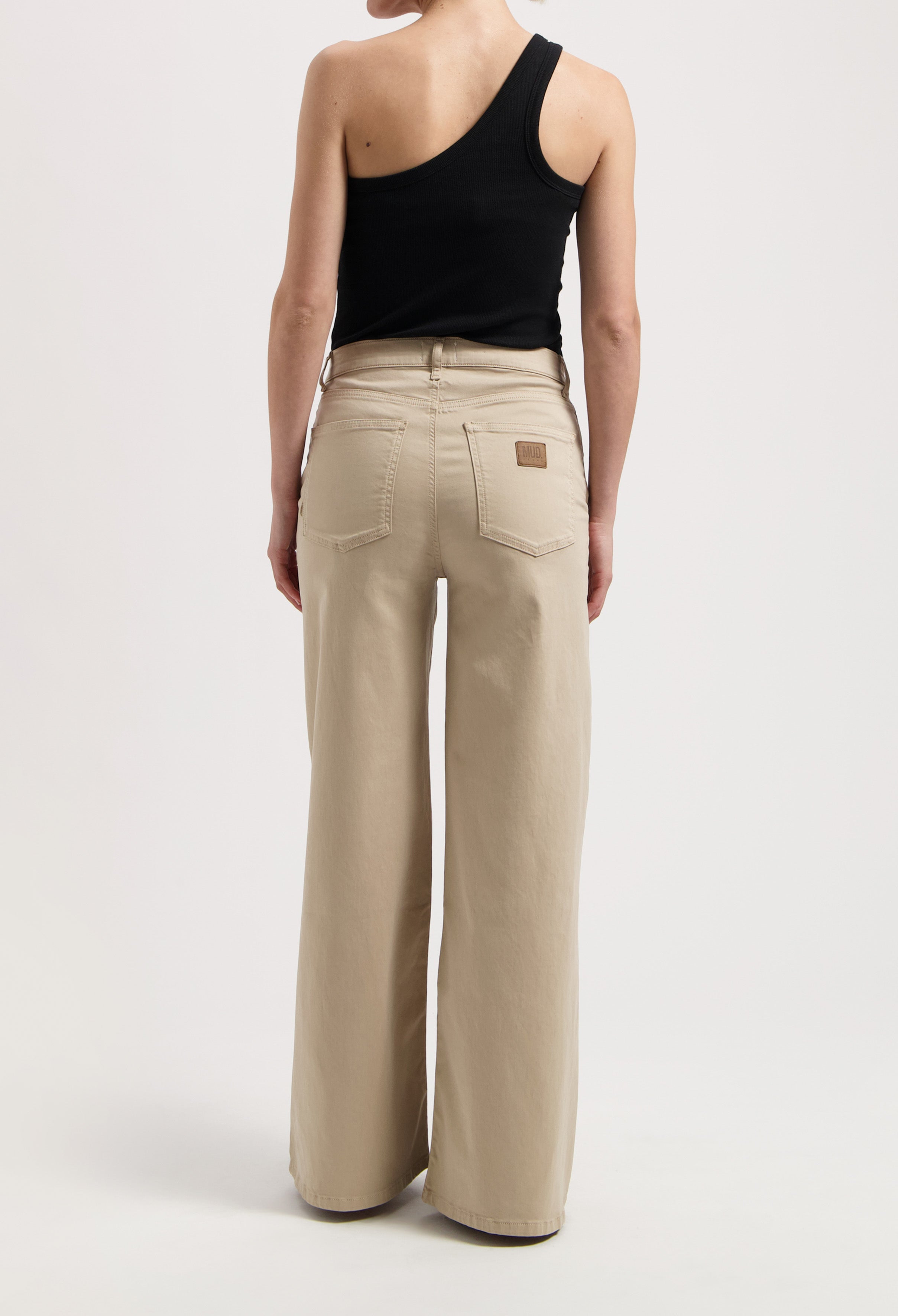 Wide-leg beige MUD Jeans made from organic cotton, worn with a black, one-shoulder sleeveless top in a minimalist style.