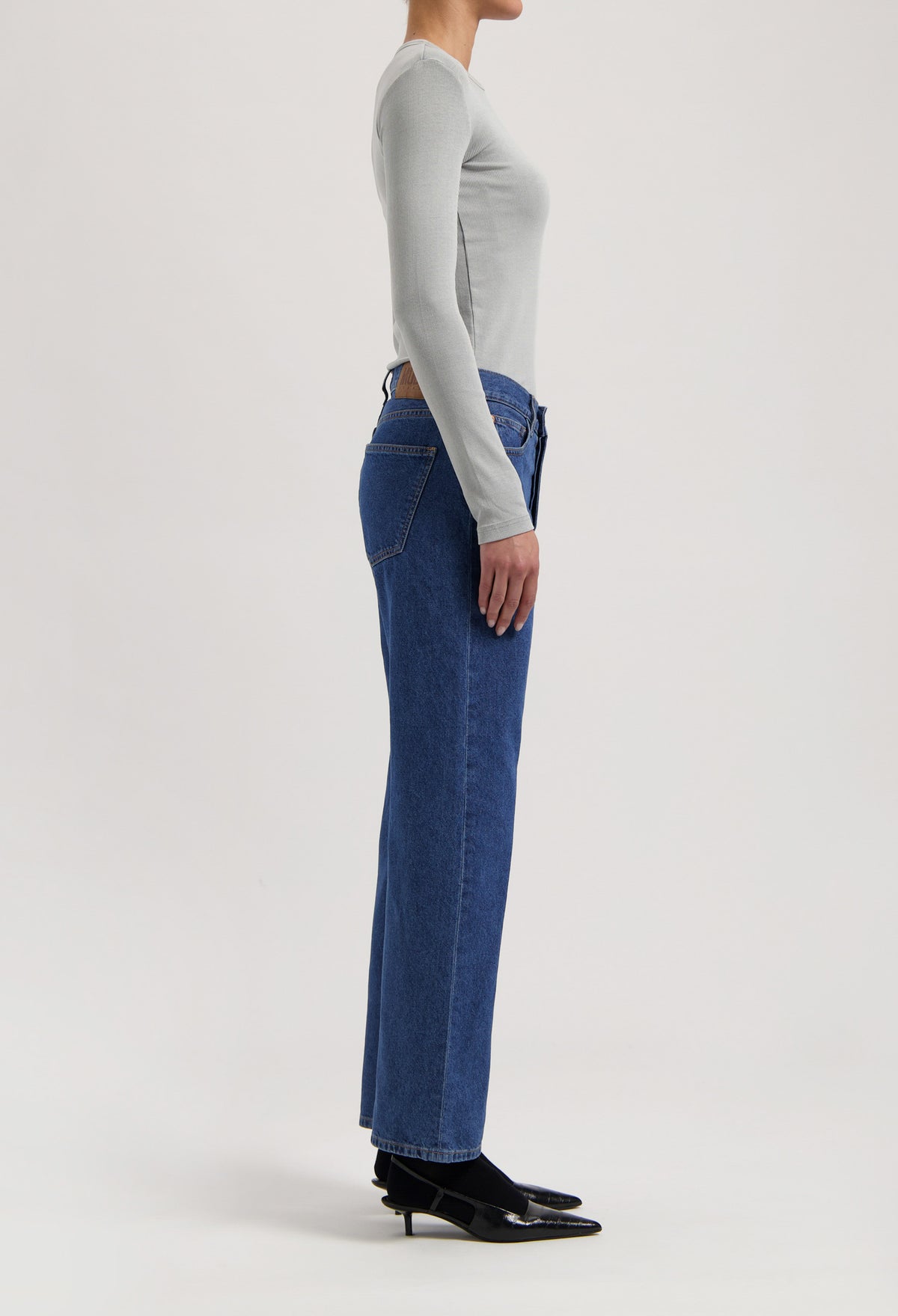 Side view of woman wearing high-waisted blue denim jeans and a light gray long-sleeve top, paired with black heels.