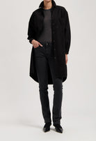 Model wearing a long black button-up coat over a gray top, paired with fitted black jeans and pointed black shoes.