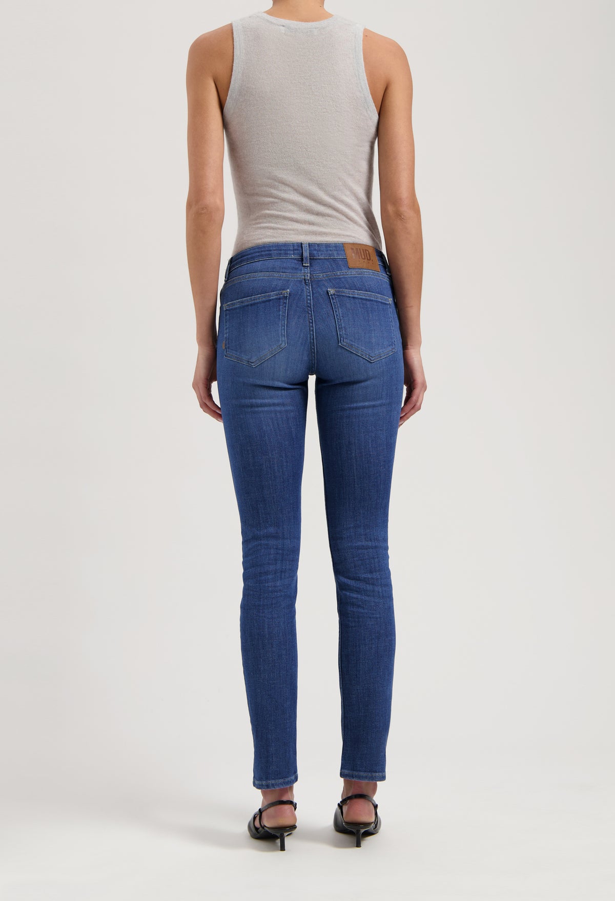 Back view of slim-fit, sustainable blue jeans by MUD Jeans, paired with a light gray sleeveless top and black sandals.