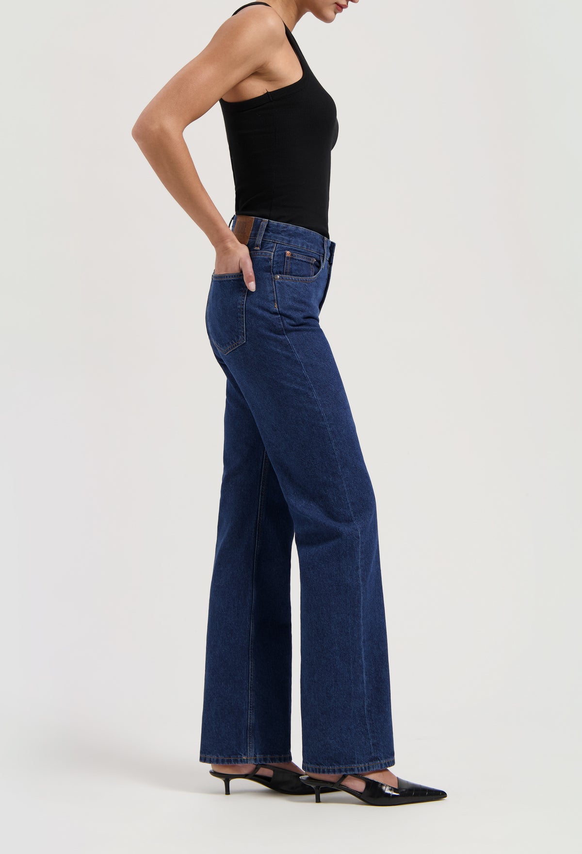 Model wearing high-waisted indigo denim jeans with a black sleeveless top and black heeled shoes on a neutral background.