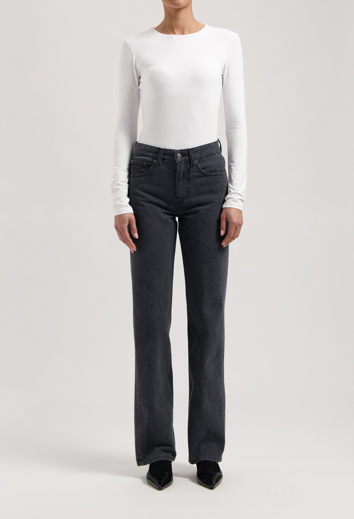 Model wearing high-waisted, black denim jeans paired with a white long-sleeve fitted top and black pointed shoes.