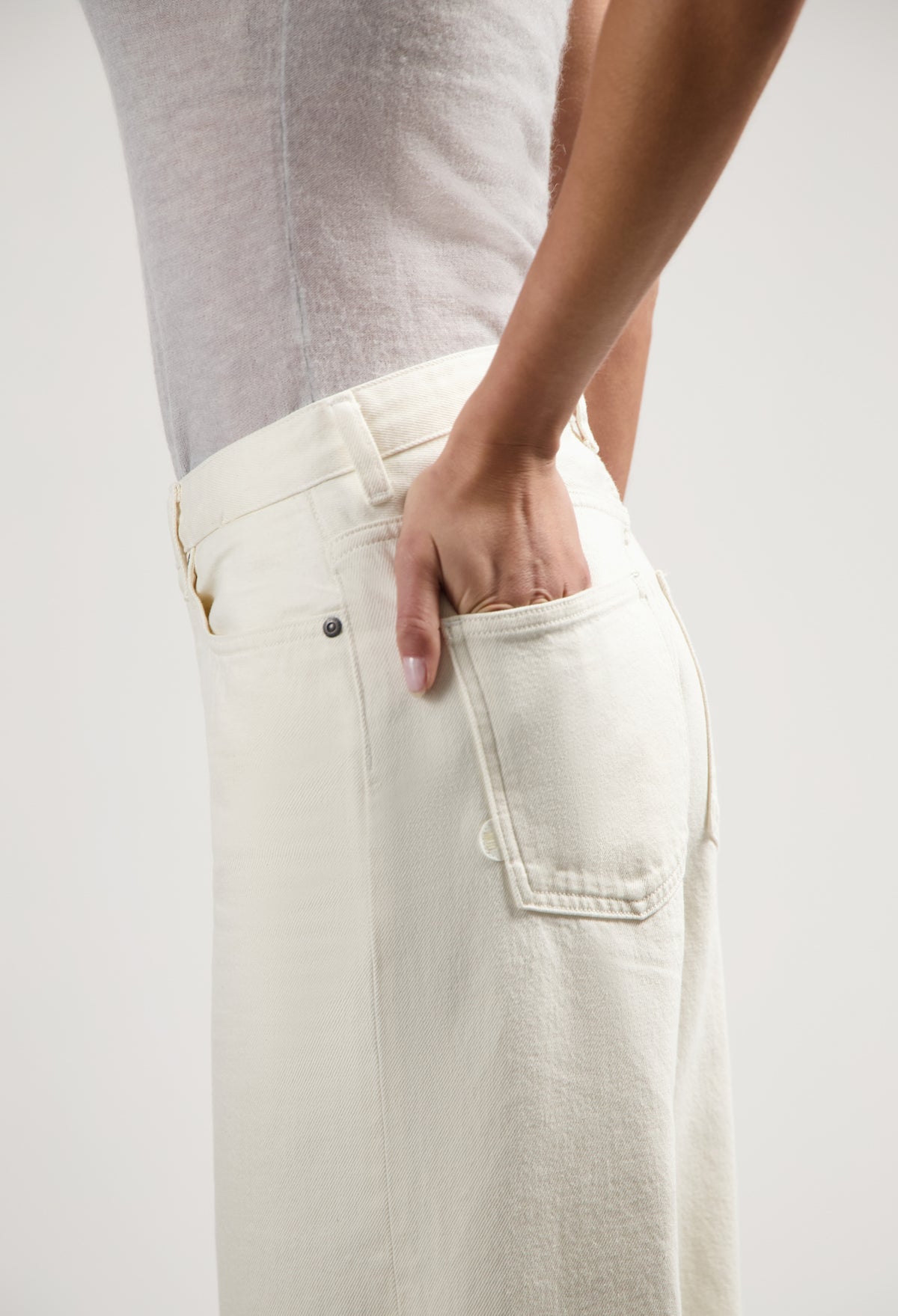 Person wearing off-white high-waisted jeans with a fitted gray sleeveless top, hand in back pocket, side view.