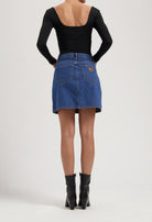 Back view of a woman in a black, long-sleeve top and a blue denim skirt, paired with sleek, black ankle boots.