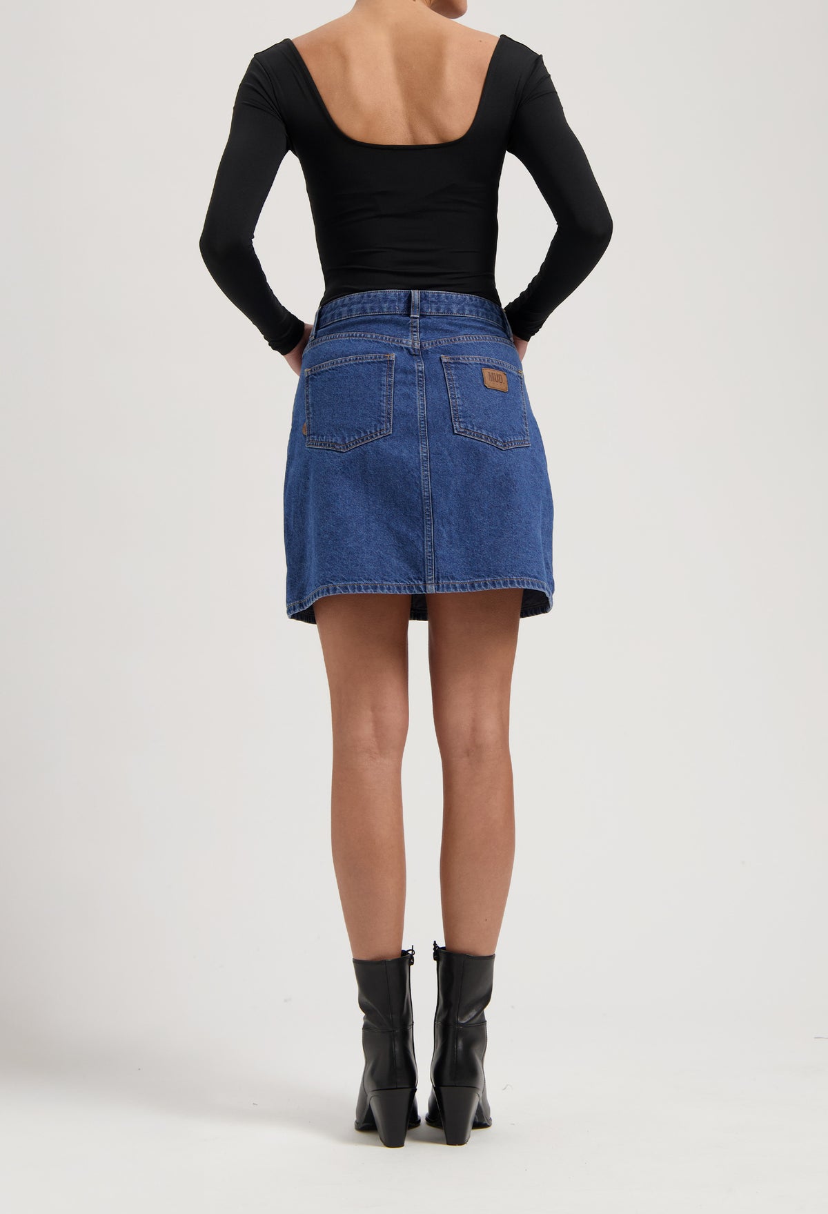Back view of a woman in a black, long-sleeve top and a blue denim skirt, paired with sleek, black ankle boots.
