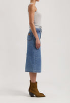 Side view of a woman in a sleeveless gray top, wearing a blue denim midi skirt paired with brown suede ankle boots.