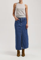 Long blue denim skirt with front slit, paired with a sleeveless gray top and brown pointy boots, showcasing casual chic style.