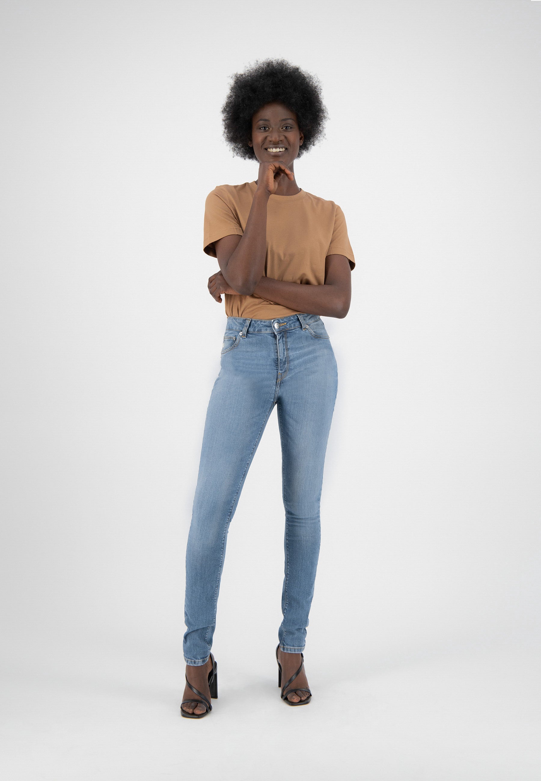 Woman modeling light blue denim jeans and tan t-shirt, wearing black heels against plain white background, smiling confidently.